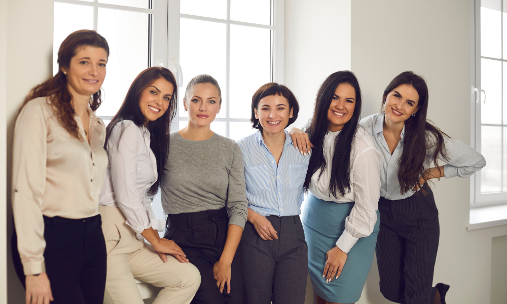 Sechs berufstätige Frauen in Businesskleidung lächeln und posieren locker in einem hellen Büro vor großen Fenstern.