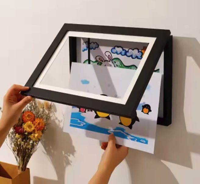 Wandrahmen mit aufklappbarer Front, Hände schieben ein Kinderbild mit Pinguinen hinein; getrocknete Blumen daneben.