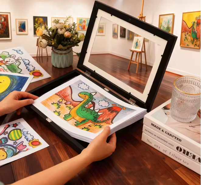 Hände legen eine bunte Kinderzeichnung eines Dinosauriers in einen schwarzen Aufklapprahmen, im Hintergrund Galerie und Blumen.