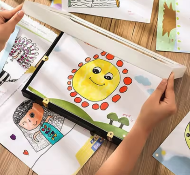 Hände öffnen eine Mappe mit einer Kinderzeichnung: lächelnde gelbe Sonne mit roten Punkten, auf einem Holztisch weitere Zeichnungen.