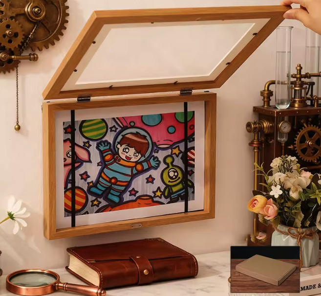 Offene Holzvitrine mit Glasdeckel zeigt ein buntes Kinderbild eines Astronauten, auf Schreibtisch neben Lederjournal und Blumen.