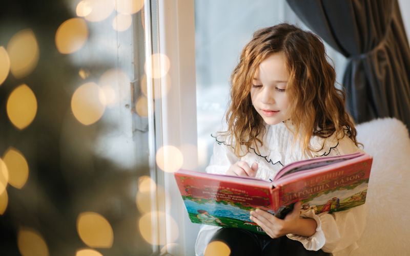 Mädchen mit lockigem Haar liest ein buntes Buch am Fenster, verschwommene Lichter im Vordergrund