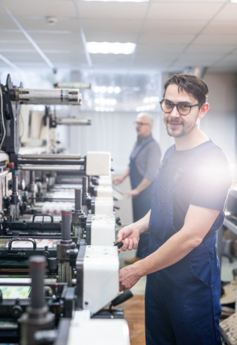 Mann in blauer Arbeitskleidung und Brille bedient Druckmaschine in Werkstatt, unscharfer Kollege im Hintergrund.